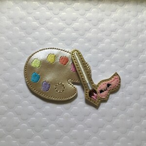 May include: A tan-colored artist's palette and paintbrush applique. The palette has colorful paint splotches in yellow, orange, green, blue, and purple. The paintbrush has a pink brush head and a brown handle.
