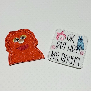 May include: Two embroidered iron-on patches. One is orange and features a fuzzy monster with a big smile. The other is white with black text that reads "Ok, but first Ms. Rachel" and features a blue jean design.