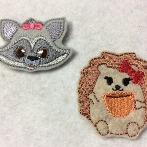 May include: Two embroidered felt patches, one of a raccoon with a pink bow and the other of a hedgehog with a red bow holding a brown cup.