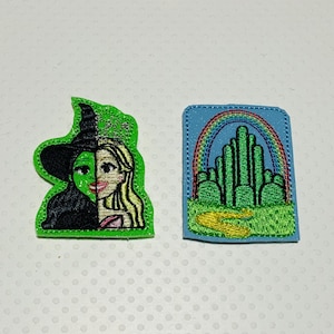 May include: Two embroidered iron-on patches. One patch is green and features a witch with green skin and a black hat. The other patch is blue and features a green emerald city with a rainbow above it.