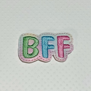 May include: A colorful embroidered patch with the letters "BFF" in green, blue, and pink. The patch has a white border and a pastel rainbow background. The letters are stitched with a textured thread.