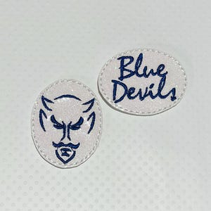 May include: Two oval-shaped patches with a glittery white background. One patch features a blue devil face, and the other says "Blue Devils" in blue script. The patches have a white border.