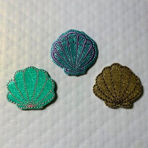 May include: Three glitter seashells in different colors: teal, purple, and gold. The shells are all the same size and shape, and they are arranged in a row.