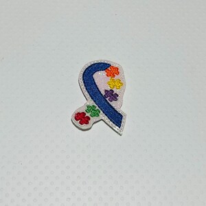 May include: Blue ribbon with a white background and rainbow puzzle pieces. The ribbon is a symbol of autism awareness.