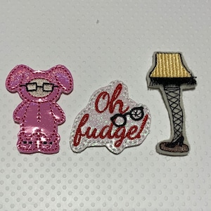 May include: Three embroidered patches featuring iconic imagery. One patch depicts a pink bunny suit with glasses, another reads "Oh fudge!" with glasses, and the third shows a leg lamp with a gold lampshade.