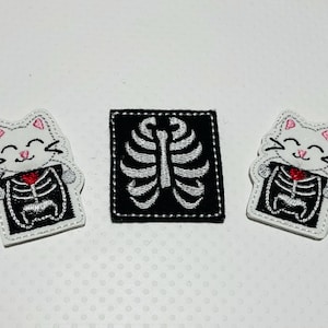 May include: Three embroidered iron-on patches featuring a white cat with a pink nose and a black skeleton inside. The cat is holding a red heart. One patch is a black square with a white skeleton inside.