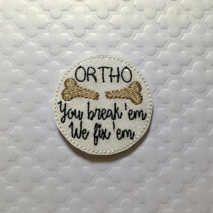 May include: White round patch with the text "ORTHO" and two gold bone shapes. The text below the bone shapes reads "You break 'em we fix 'em".