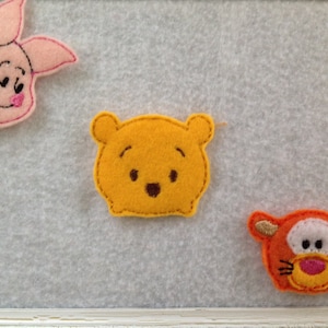 May include: Three felt appliques of cartoon characters: a pink pig with a smile, a yellow bear with a smile, and an orange tiger with a smile.
