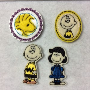 May include: Four embroidered iron-on patches featuring characters from the Peanuts comic strip. The patches are yellow, white, and blue. One patch features Snoopy, another features Charlie Brown, and the other two feature Lucy and Linus.