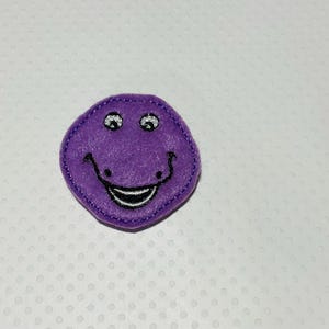 May include: A purple felt applique in the shape of a smiling face. The applique has black stitched details for the eyes, mouth, and outline. The eyes have silver glitter accents. The background is a white surface with a subtle dotted pattern.