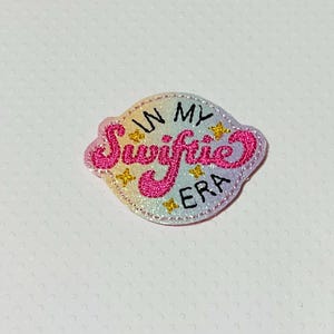 May include: A pink and white embroidered patch with the text "In My Swiftie Era" in a rainbow-colored background.