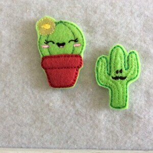 May include: Two felt cactus ornaments. One is green with a smiling face and a mustache. The other is green with a pot and a flower on top, and a smiling face.