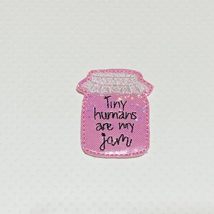 May include: A pink patch in the shape of a jar with the text "Tiny humans are my jam" in black script. The jar has a glittery outline and a white lid.