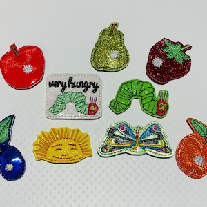 May include: A collection of colorful, glittery fabric appliques featuring fruits, a caterpillar, a butterfly, and a sun. The text "very hungry" is on one applique. The appliques include a red apple, a green pear, and a red strawberry.