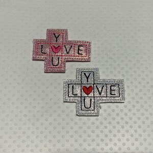 May include: Two embroidered iron-on patches in the shape of a cross. The top patch is pink with the words "LOVE YOU" and a heart in the center. The bottom patch is silver with the words "LOVE YOU" and a heart in the center.