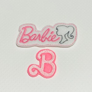May include: Two pink felt patches with embroidered designs. One patch says "Barbie" with a black outline of a woman's head. The other patch is a pink letter "B" in a cursive font.