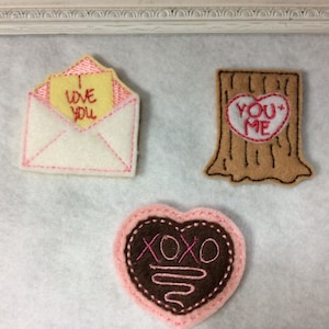 May include: Three felt Valentine's Day decorations. An envelope with "I LOVE YOU" on a yellow note, a tree stump with a heart that says "YOU + ME", and a pink heart with "XOXO" on it.