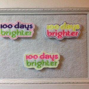 May include: Three white felt patches with green, pink, and yellow embroidery. Each patch reads "100 days brighter".