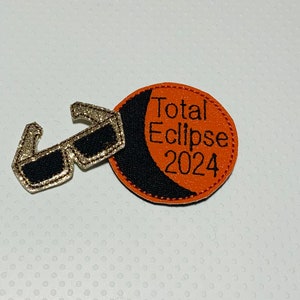 May include: Orange and black felt patch with the text "Total Eclipse 2024" and a pair of silver glitter sunglasses.
