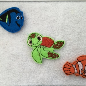 May include: Three felt appliques of characters from the movie Finding Nemo. A blue and black Dory, a green turtle with a red shell, and an orange and white clownfish.