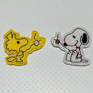 May include: Two embroidered patches featuring cartoon characters. One is yellow with a black outline, holding a small object. The other is white with black ears, a red collar, and is holding a brown object. Both have glittery accents.