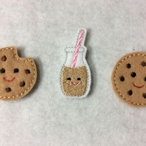 May include: Three felt appliques with a smiling face design. A brown chocolate chip cookie, a brown chocolate chip cookie with a pink straw, and a brown milk bottle with a pink straw.