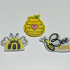 May include: Three embroidered iron-on patches with a bee theme. The first patch is a yellow and black bee with a rainbow and a heart. The second patch is a yellow and black bee with the words "Bee Kind". The third patch is a yellow and black beehive with a heart.