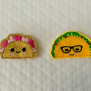 May include: Two felt taco appliques with embroidered faces. One taco is yellow with green trim and glasses. The other taco is gold with pink hearts and glitter.