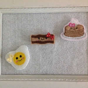 May include: Three felt appliques in the shape of breakfast foods. A brown and white bacon strip with a red bow, a yellow and white fried egg with a yellow bow, and a stack of brown pancakes with a pink bow and white frosting.