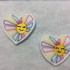 May include: Two embroidered felt heart-shaped patches with a yellow smiling sun face in the center. The sun face has pink cheeks and closed eyes. The sun face is surrounded by white, pink, blue, and purple rays.