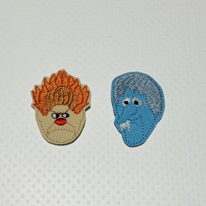 May include: Two embroidered patches featuring cartoon faces. One has a tan face with orange hair, a red mouth, and black eyes. The other has a blue face with silver hair and white accents. Both are on a white dotted background.