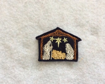 May include: An embroidered patch depicting a nativity scene with a brown wooden stable, a dark blue interior, two white figures, and three yellow stars.