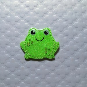 May include: A green glitter frog applique with embroidered eyes, a smile, and spots.