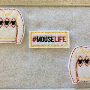 May include: White felt sweater patches with a V-neck and a diamond pattern, featuring black Mickey Mouse heads. A rectangular patch with the text "#MOUSELIFE" is also present. The patches are outlined in red and yellow.
