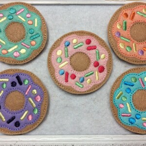 May include: Five felt donut-shaped coasters with colorful frosting and sprinkles. The coasters feature light brown bases with various frosting colors, including green, pink, orange, purple, and blue. Each coaster has a stitched border and colorful sprinkles.