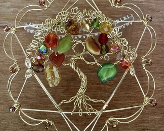 May include: A wire-wrapped tree sculpture crafted from gold-toned wire, featuring a central tree design with multi-colored glass beads in shades of green, amber, and red. The tree is enclosed within a geometric hexagonal wire frame.