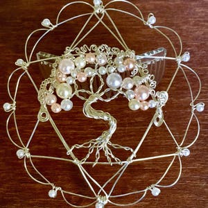 May include: A silver wire tree of life hair comb with pink and white pearls. The comb is set in a silver wire circle with a geometric design.