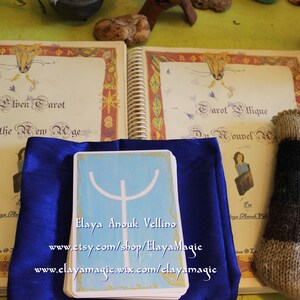 May include: A blue cloth covers a tarot card with a white symbol on a light blue background. The card reads "Elaya Anouk Vellino" and includes the website addresses "www.etsy.com/shop/ElayaMagic" and "www.elayamagic.wix.com/clayamagic". The card is next to a book titled "Elven Tarot of the New Age" and a book titled "Tarot &Eacute;thique Du Nouvel Age".
