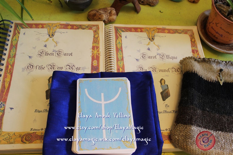 May include: A blue cloth covers a tarot card with a white symbol on a light blue background. The card reads "Elaya Anouk Vellino" and includes the website addresses "www.etsy.com/shop/ElayaMagic" and "www.elayamagic.wix.com/clayamagic". The card is next to a book titled "Elven Tarot of the New Age" and a book titled "Tarot &Eacute;thique Du Nouvel Age".