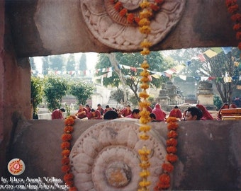 May include: A group of people wearing red robes are seated in front of a stone structure adorned with intricate carvings and yellow flower garlands. The structure is partially obscured by a stone wall with a window opening. The image is taken from inside the structure, looking out through the window.