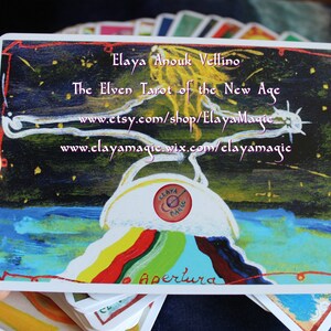 May include: A tarot card with a colorful rainbow design and the text "Apertura" at the bottom. The card is from the "The Elven Tarot of the New Age" deck by Elaya Anouk Vellino. The card features a white circle with the text "Claya Magic" in the center.