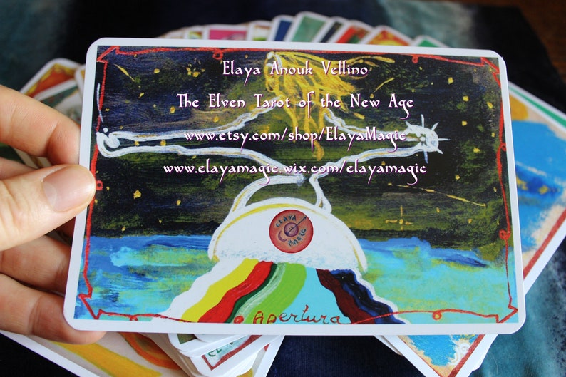 May include: A tarot card with a colorful rainbow design and the text "Apertura" at the bottom. The card is from the "The Elven Tarot of the New Age" deck by Elaya Anouk Vellino. The card features a white circle with the text "Claya Magic" in the center.