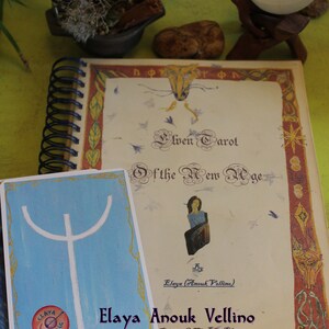 May include: A closed book with a colorful cover and the title "Elven Tarot of the New Age" by Elaya (Anouk Vellino). The book is on a table with a white candle and a small cauldron.