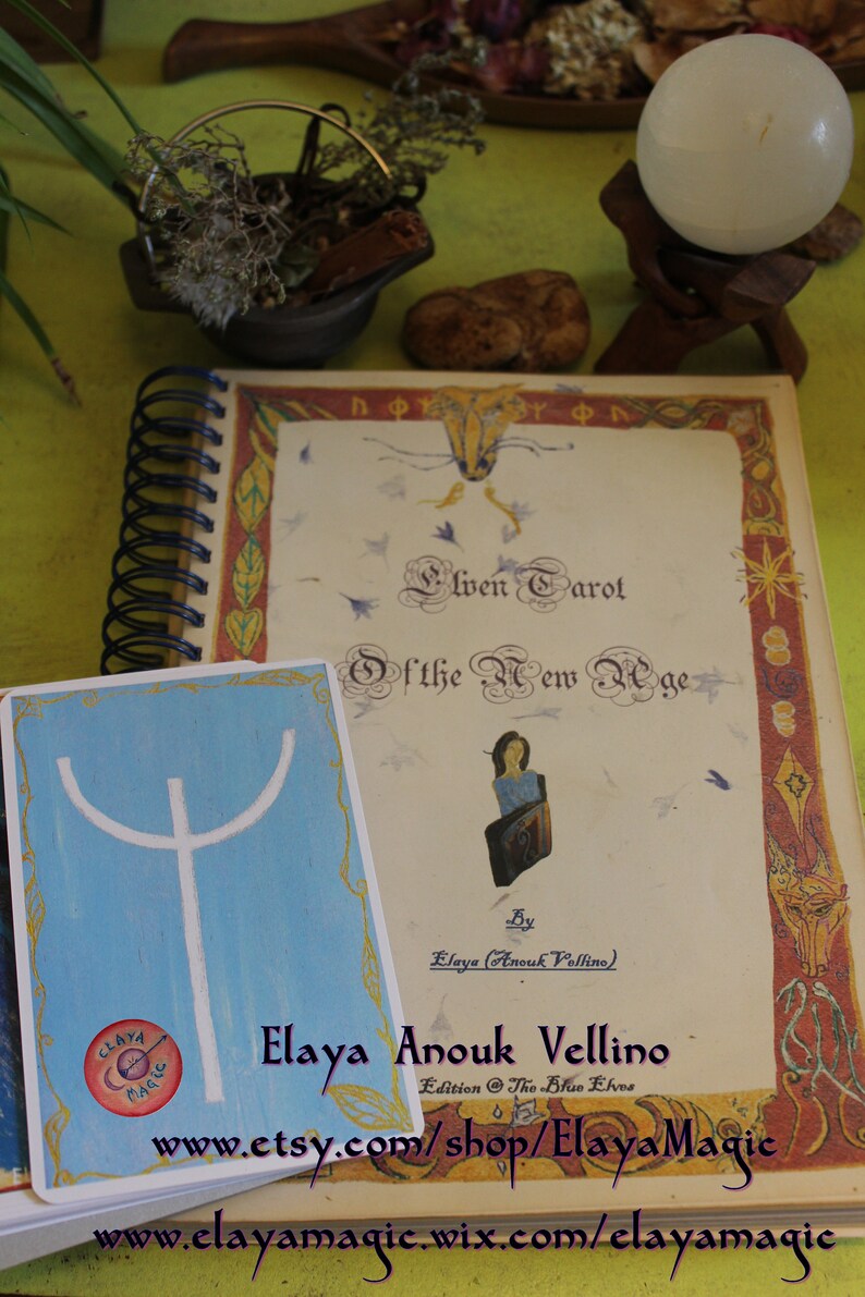 May include: A closed book with a colorful cover and the title "Elven Tarot of the New Age" by Elaya (Anouk Vellino). The book is on a table with a white candle and a small cauldron.