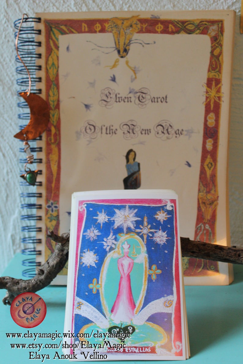 May include: A close-up of a tarot card from the Elven Tarot of the New Age deck. The card depicts a woman in a pink dress standing in a circle of stars against a blue background. The card is titled "Estrellas" and features the text "Elven Tarot of the New Age" and "Elaya Anouk Vellino".