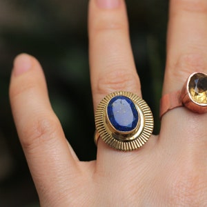Könnte beinhalten: Zwei Ringe mit Edelsteinen. Der Ring links hat einen blauen ovalen Stein, der in einem goldenen Band mit einem Sonnenstrahl-Design gefasst ist. Der Ring rechts hat einen gelben ovalen Stein, der in einem Kupferband gefasst ist.