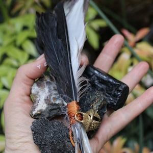 Puede incluir: Una mano sosteniendo una pluma negra y blanca con una cuerda naranja y un amuleto de campana dorada. La pluma está atada a un trozo de piedra negra y blanca.