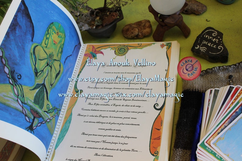 May include: An open book with a yellow and orange cover. The book has a handwritten inscription in French. The book is open to a page with a drawing of a woman with green skin and a blue background. The book is surrounded by other items, including a small potted plant, a stone with the words "inner peace" written on it, a small round pin with the words "Claya Magic" on it, and a deck of tarot cards.