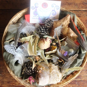 Può includere: Un cesto di vimini pieno di elementi naturali, tra cui piume, pigne, foglie secche e una piccola borsa di stoffa. Il cesto è su una superficie di legno. Il testo sulla carta dice "www.etsy.com/shop/ElwynMagic".