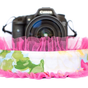 May include: A black Canon camera with a lens is partially visible behind a floral patterned camera strap. The strap features a light blue and white floral design with green accents, and is trimmed with bright pink tulle.